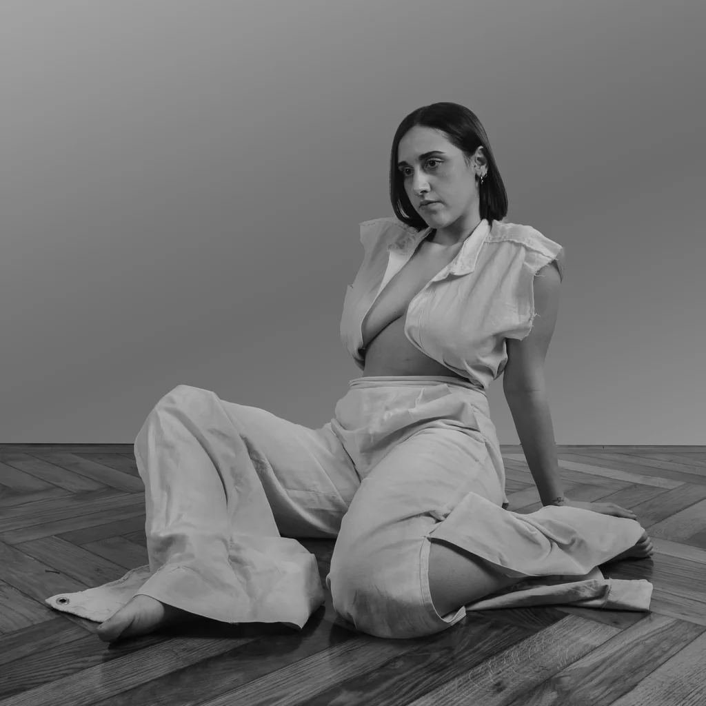 Seated on a parquet floor in a white linen shirt and wide trousers