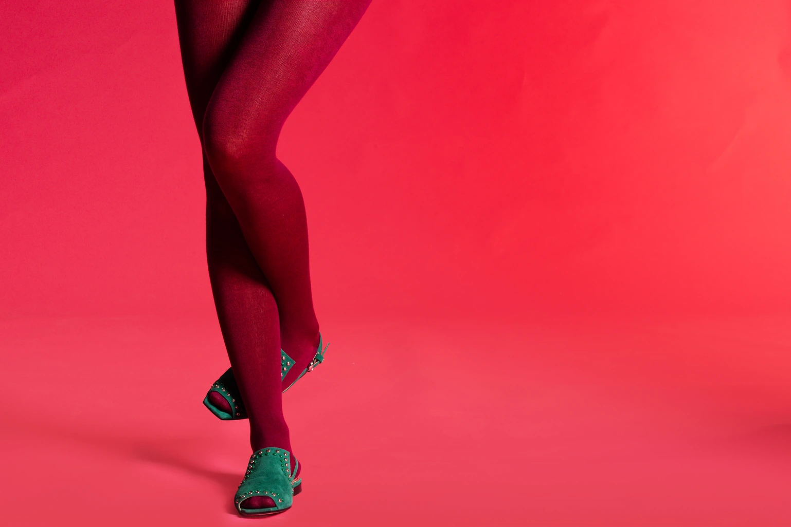 Legs in burgundy tights and teal studded sandals on a red backdrop