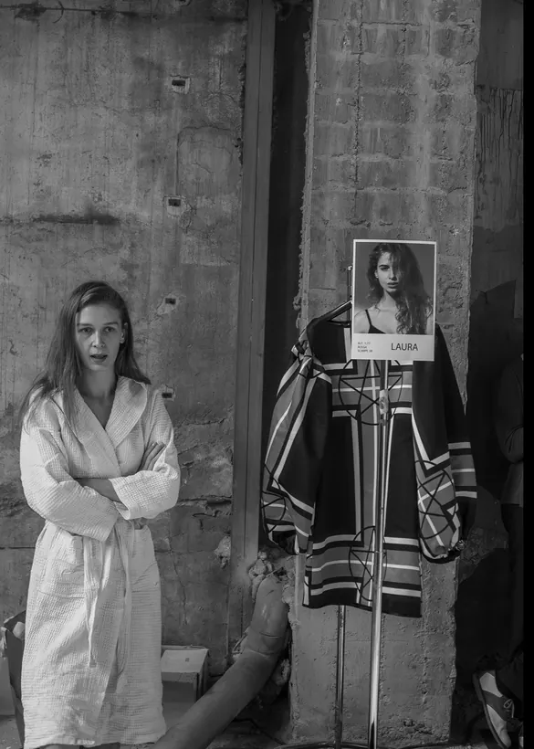 Model in a white robe backstage beside a rail with her casting card